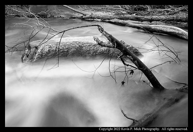 BW photograph of fallen trees and a boulder in a waterway.