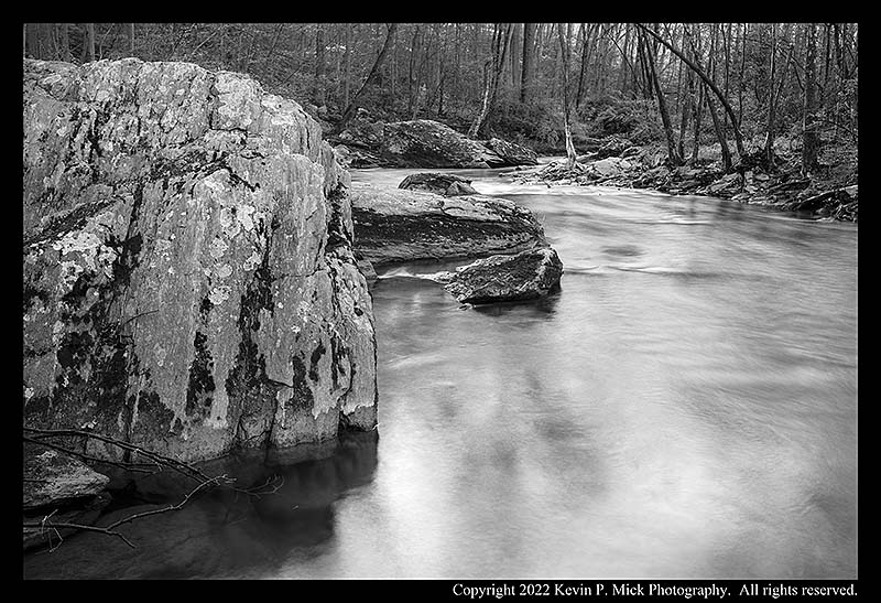 BW photograph of water flowing downstream around a large boulder.