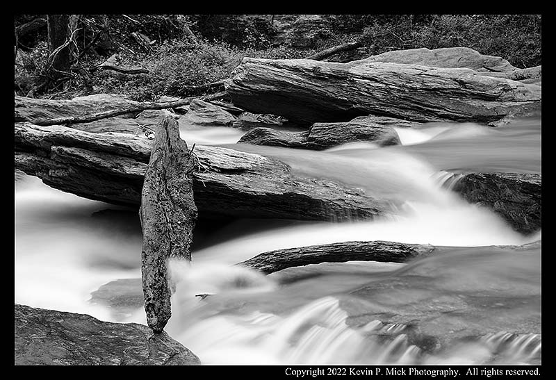 BW photograph of tree debris caught amid boulders in fast running water.